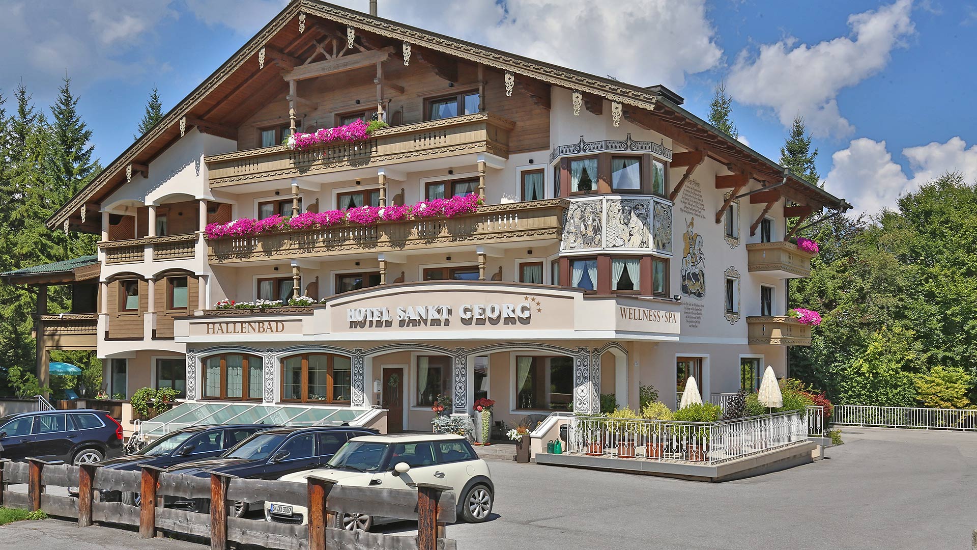 Hotel St. Georg - Alpenhotel mit Holzfassade, Balkonen voller Blumen, geparkten Autos und Restaurantterrasse bei sonnigem Wetter.