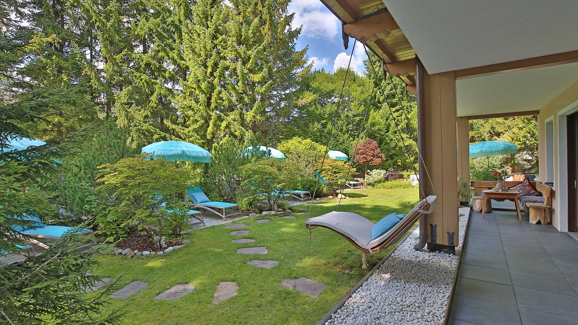 Gartenansicht mit Rasen, Gehwegplatten, Liegestühlen und türkisfarbenen Sonnenschirmen neben einer überdachten Terrasse.