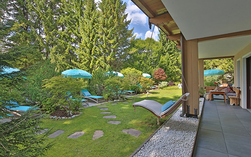 Gartenansicht mit Rasen, Gehwegplatten, Liegestühlen und türkisfarbenen Sonnenschirmen neben einer überdachten Terrasse.